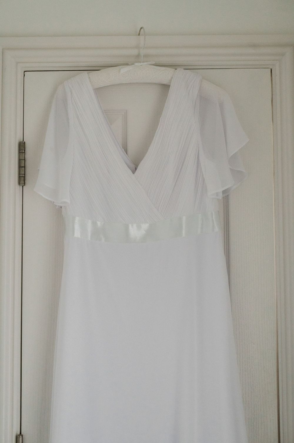 A white wedding dress with flutter sleeves and empire waist hanging on a white door.
