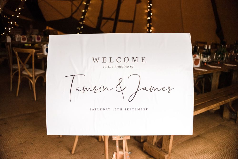 Elegant welcome sign for Tamsin and James wedding displayed on wooden easel with fairy lights in background.