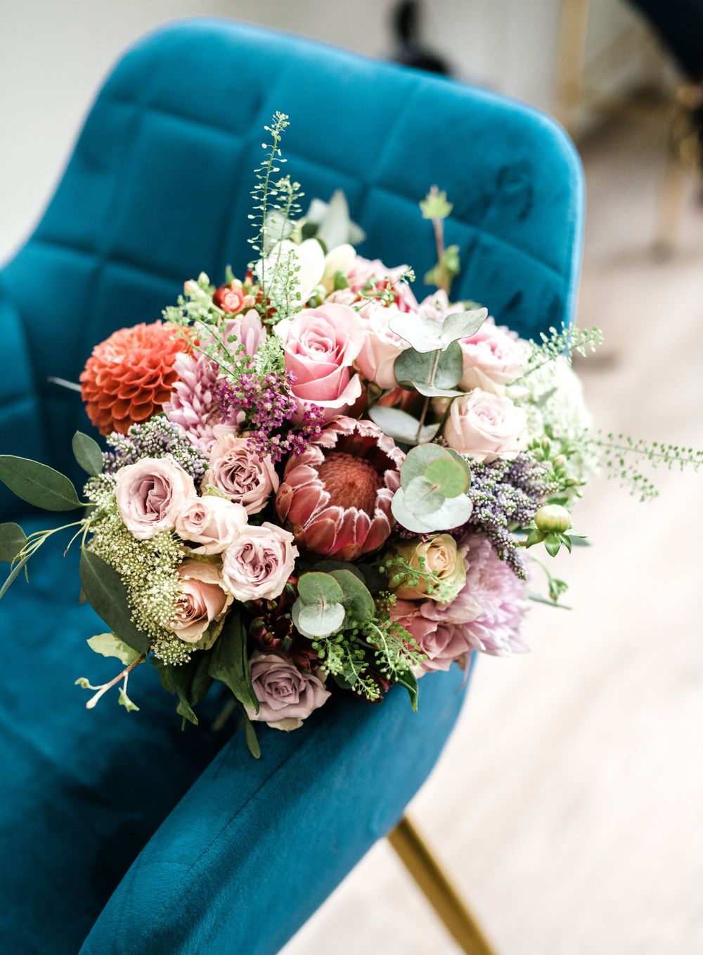 A lush bridal bouquet with pink roses and coral flowers rests on a teal velvet chair.