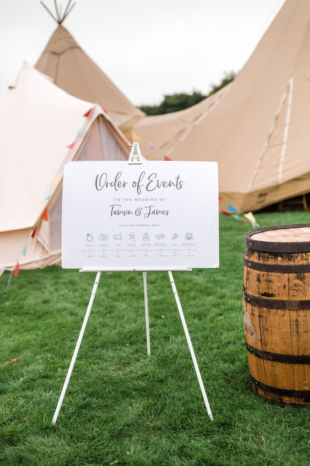 Wedding order of events sign displayed on easel with rustic barrel and teepee tents in background.