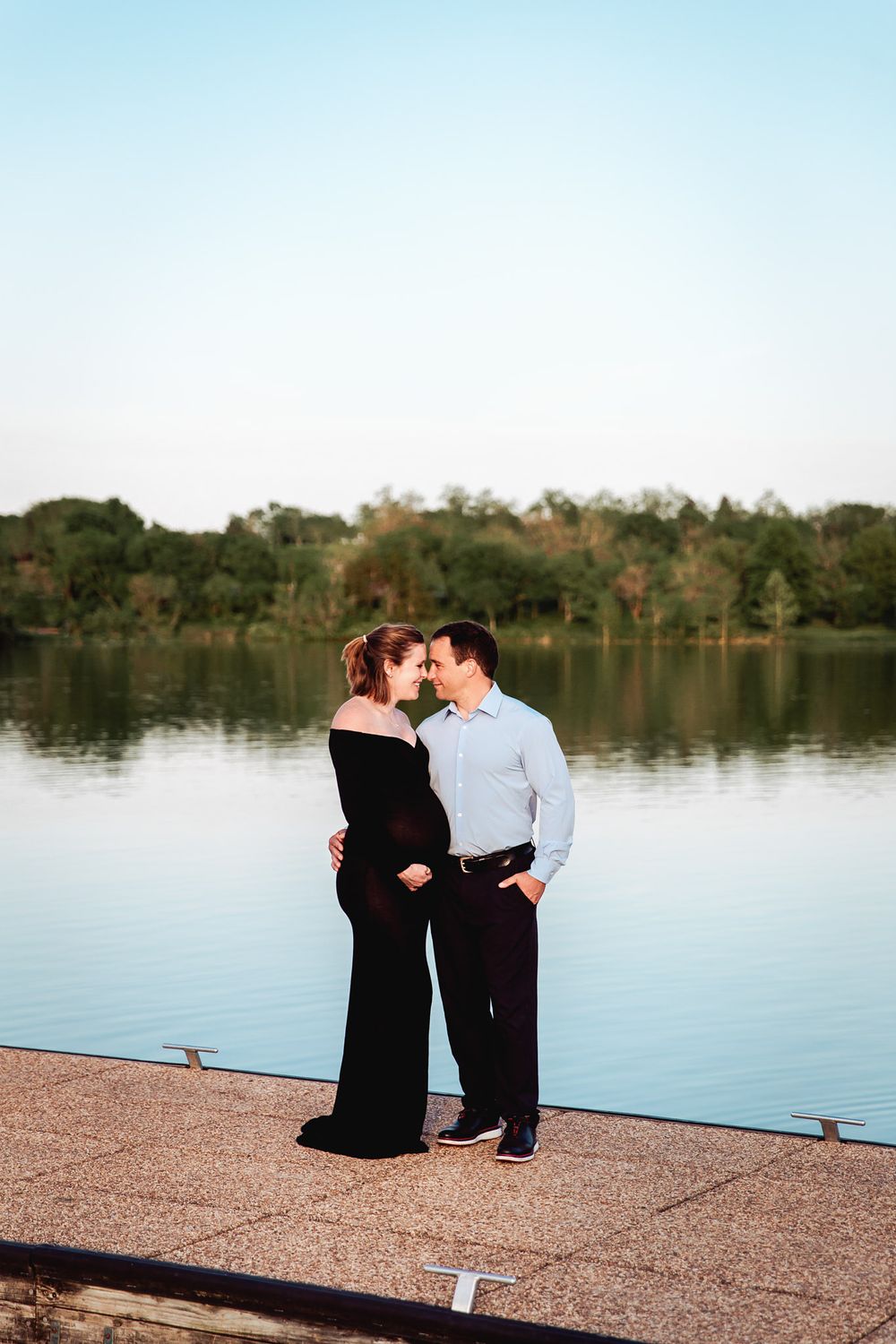 expecting mother and her husband posed on a dock with smooth lake water in background