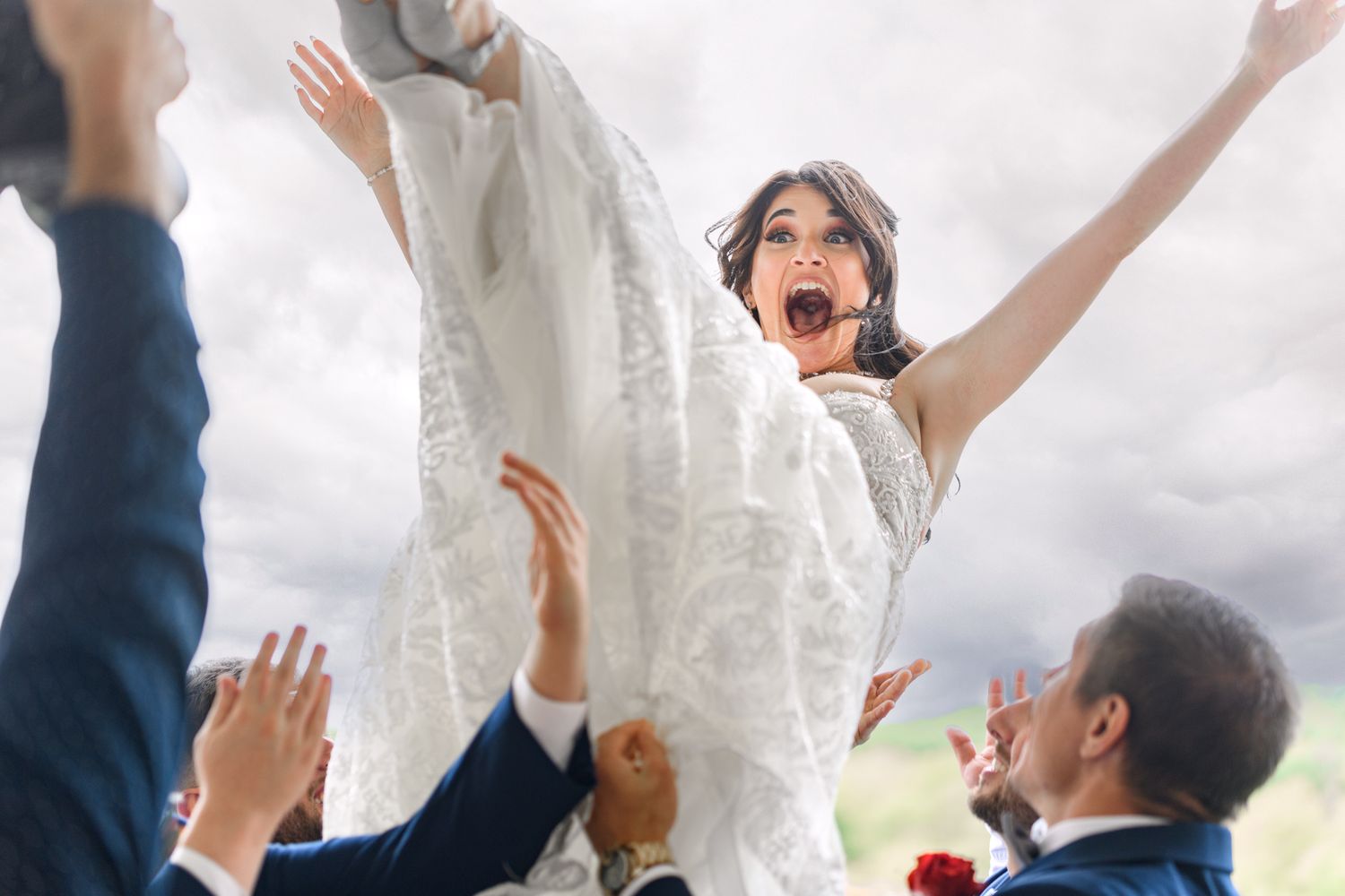 A joyous celebration with the newlyweds being lifted by guests against a cloudy sky.