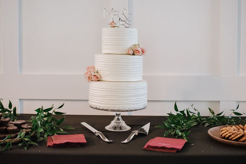 Three-tiered white wedding cake with pink floral accents and cake cutting set on dark table with greenery.