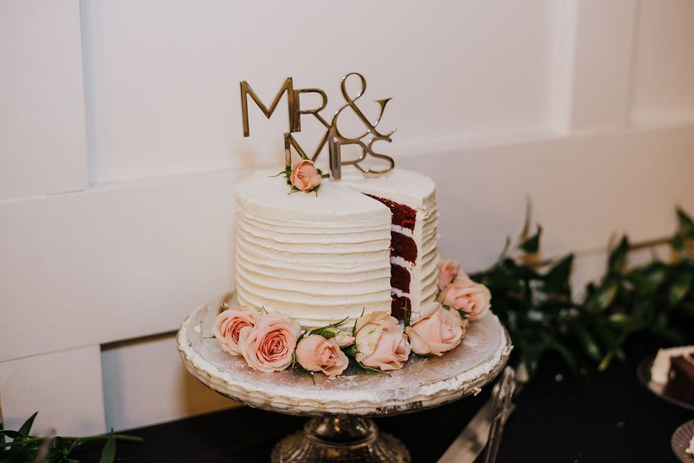 White wedding cake decorated with peach roses and gold Mr & Mrs topper on elegant cake stand.