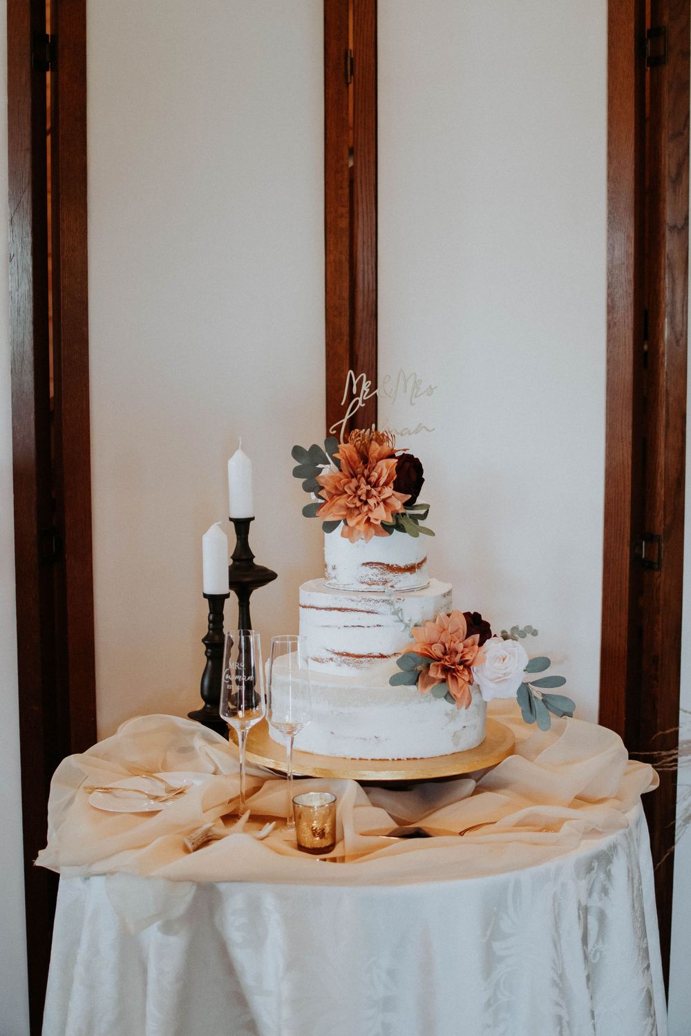 Three-tier white wedding cake with orange flowers and greenery accents on draped table.