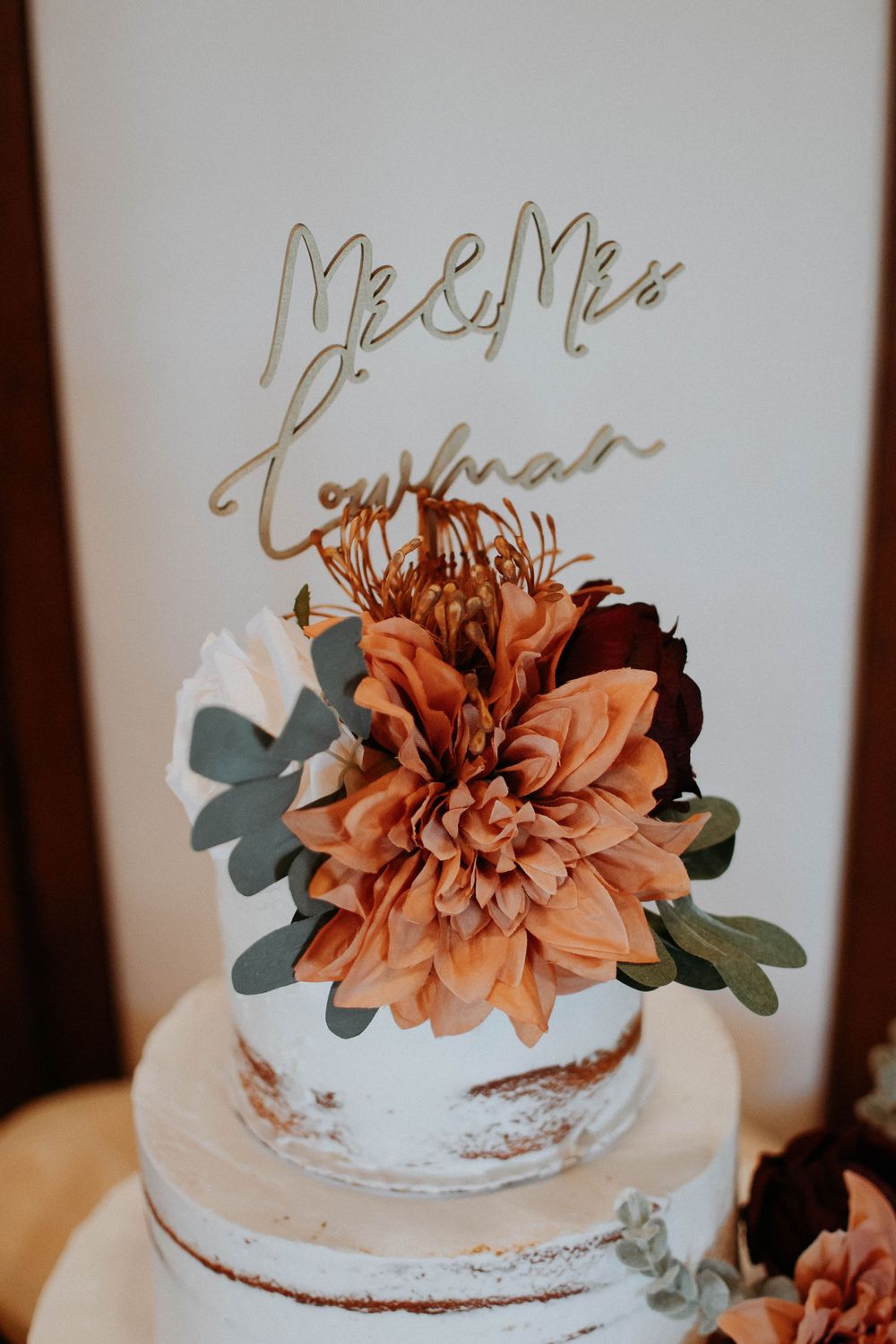 Semi-naked wedding cake topped with large orange dahlia and gold Mr & Mrs script topper.