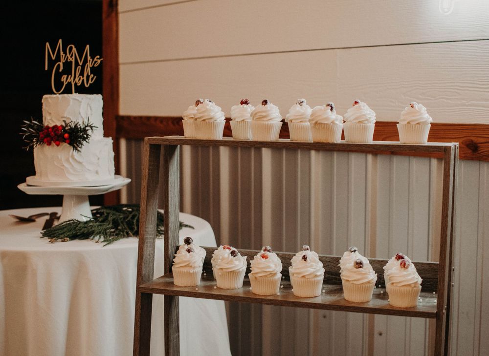 Elegant white wedding cake display with cupcakes arranged on wooden tiered shelves at a rustic reception.
