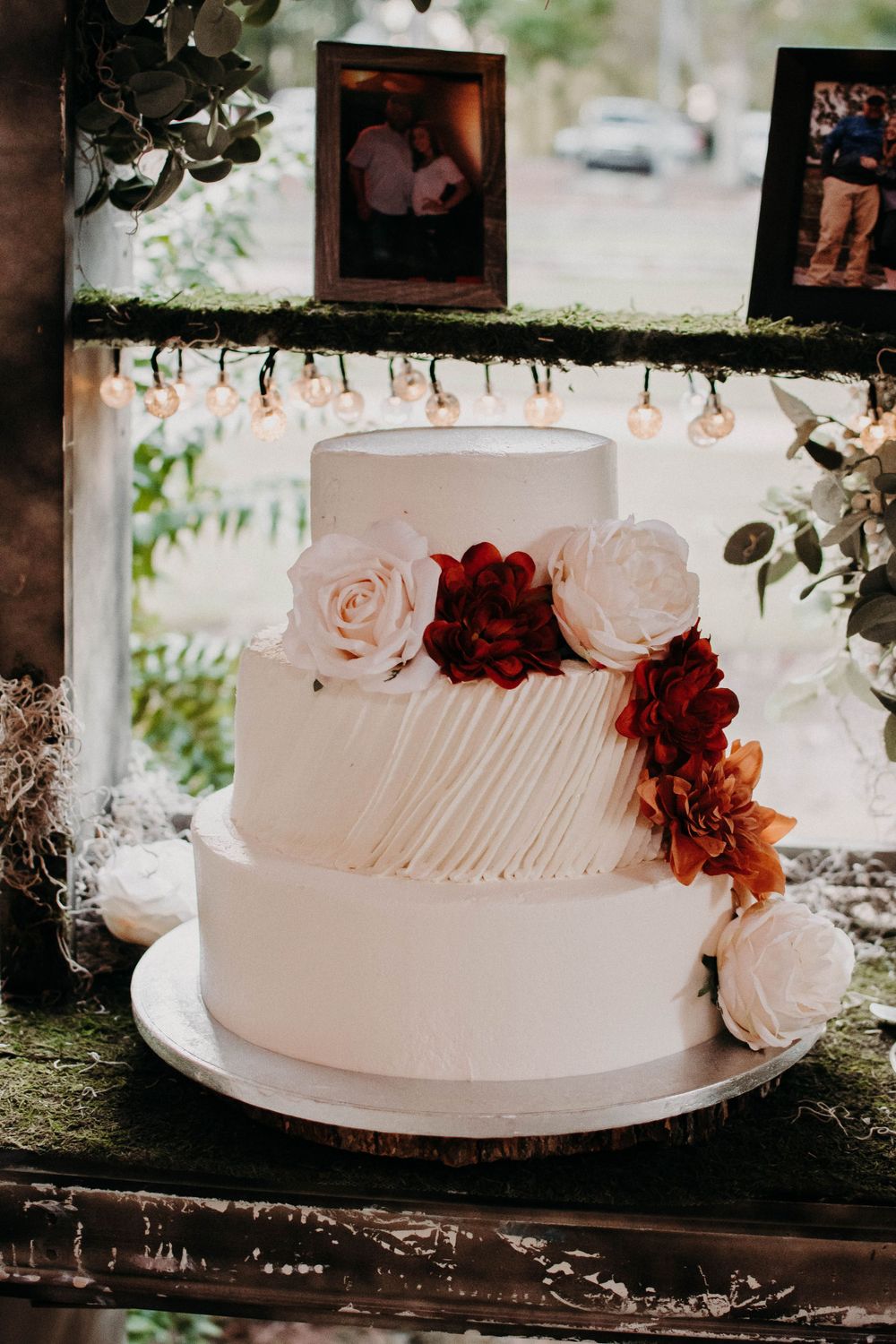 Elegant white wedding cake with textured frosting and burgundy floral decorations displayed on rustic shelf.