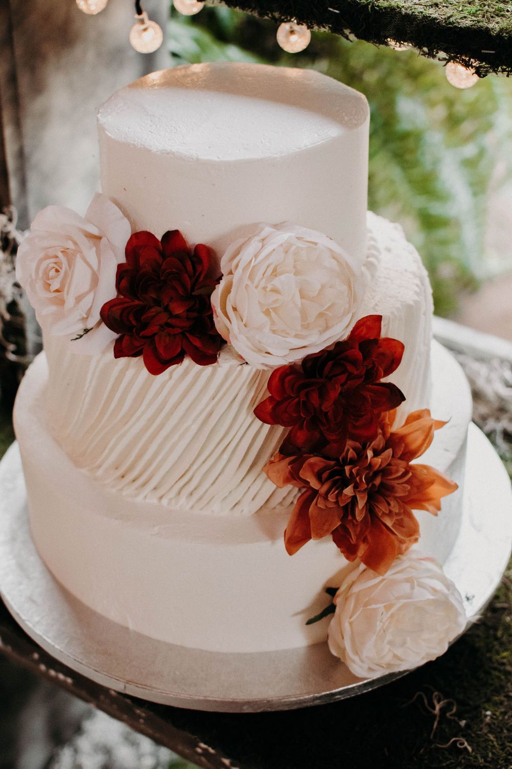 White tiered wedding cake adorned with burgundy and blush flowers in a romantic garden setting.