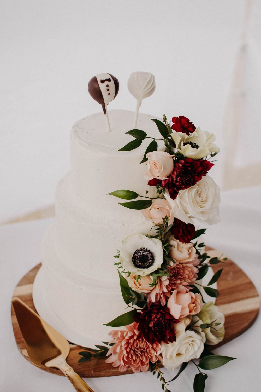 White tiered wedding cake decorated with burgundy and blush florals on wooden cake stand.