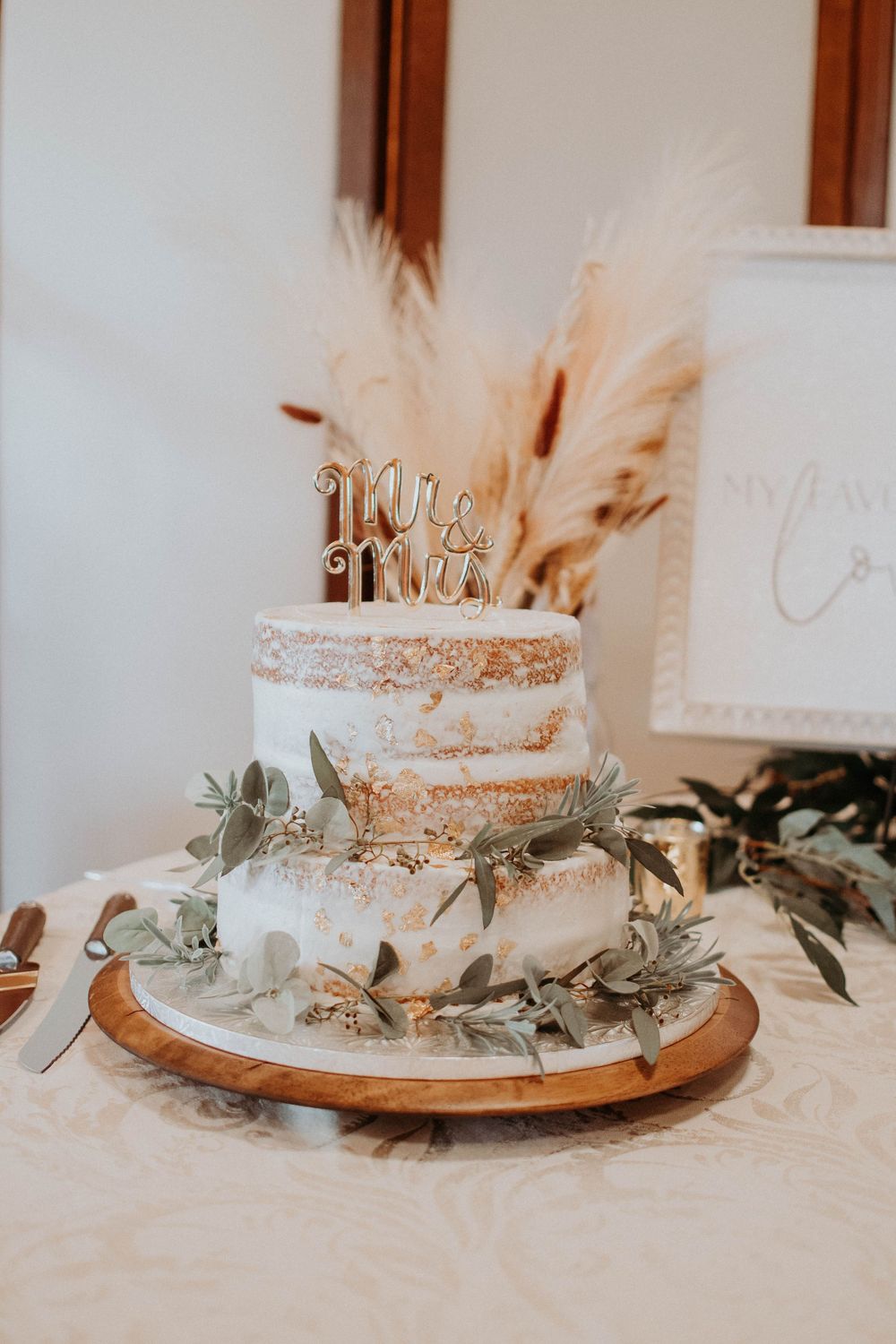 Semi-naked wedding cake decorated with pampas grass and greenery on a wooden cake stand.