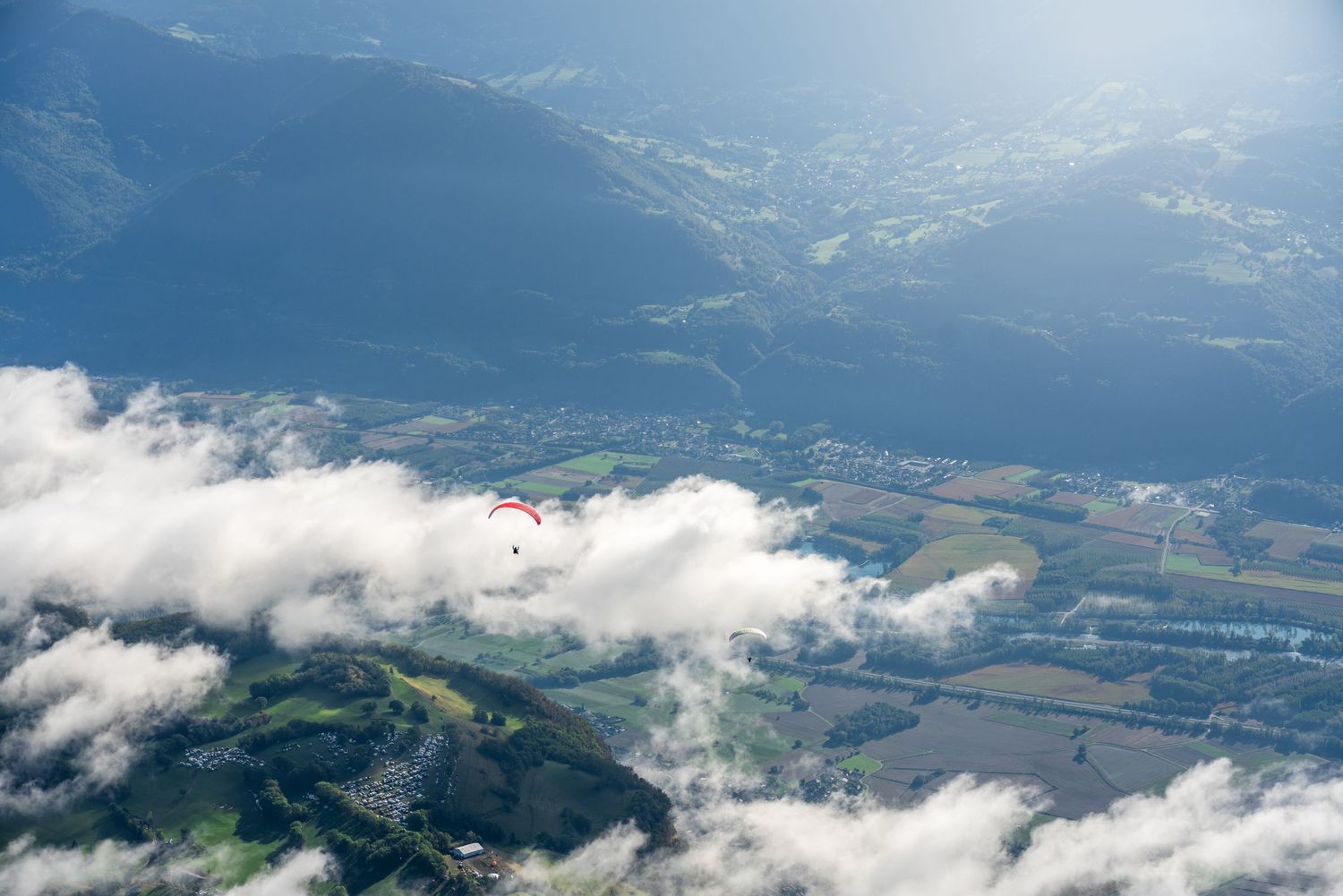 Paraglider visible through clouds above mountain valley and farmland.