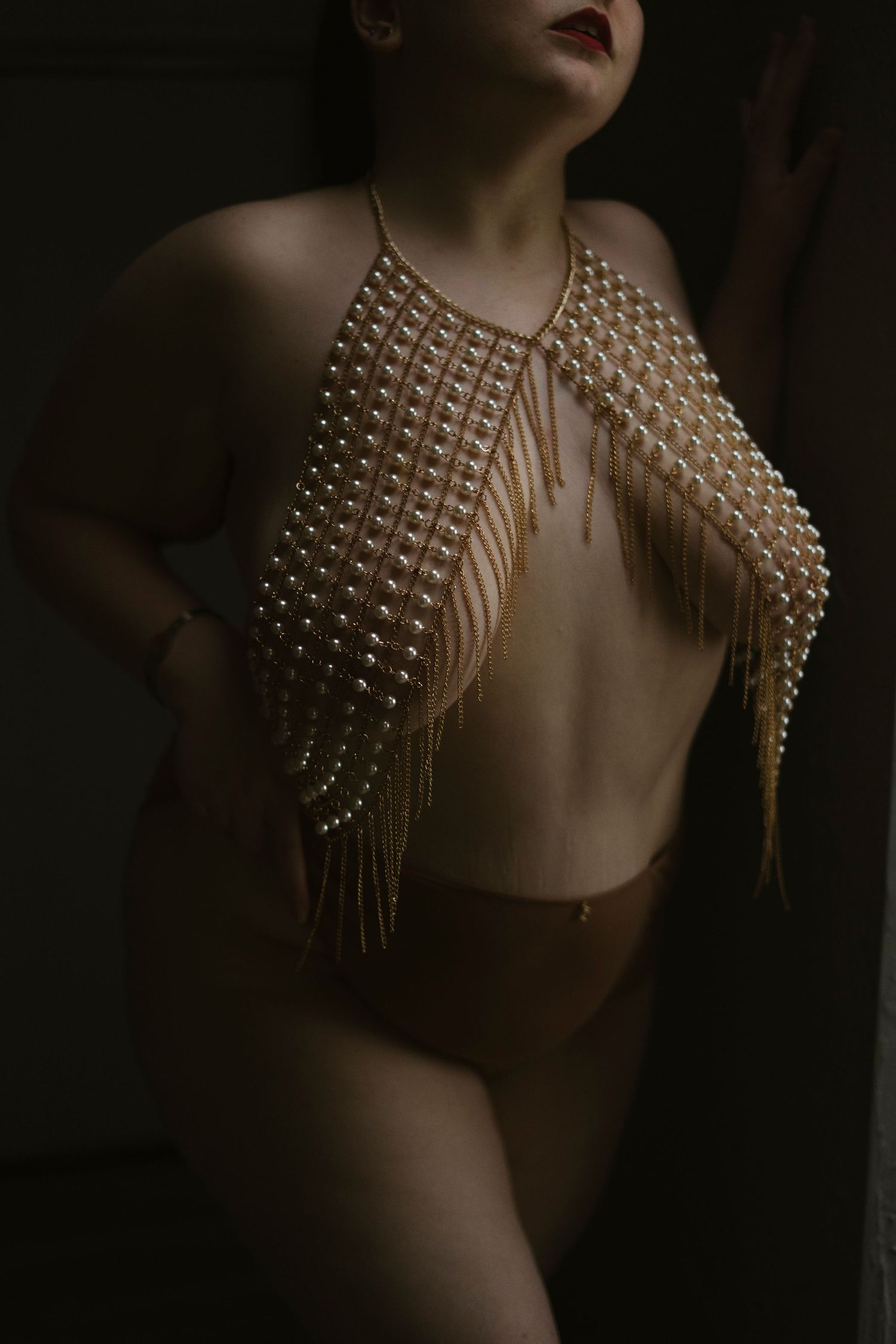moody photo of a person with bold red lipstick and a pearl and gold body chain covering their breasts