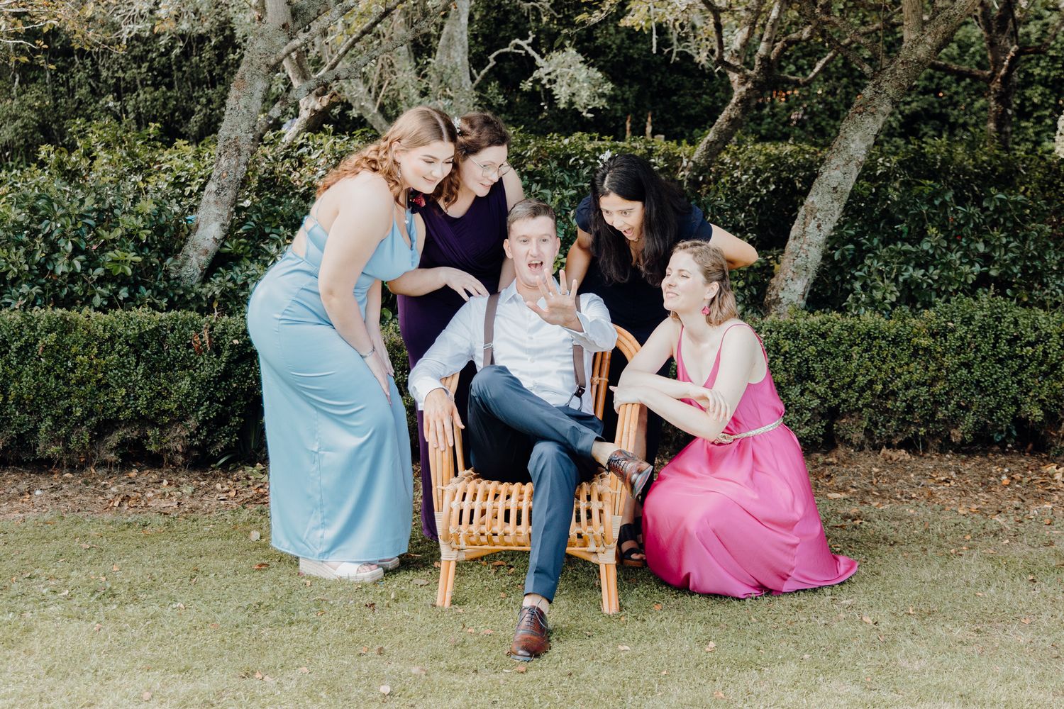 Wedding guests in pastel dresses gather around a wooden bench in a garden setting.