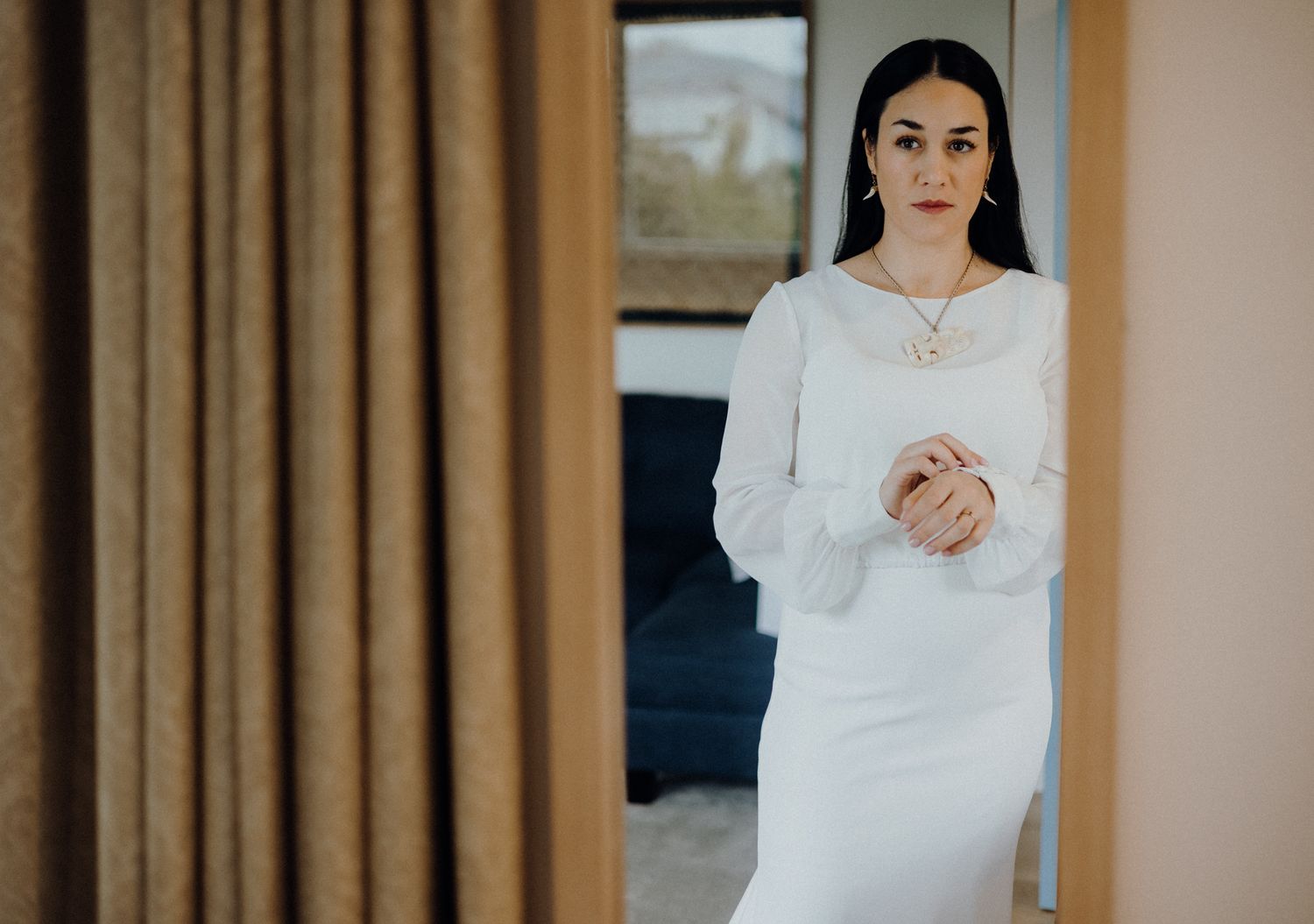 Woman in long white wedding dress standing elegantly against curtained window.