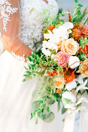 bride in lace sleeves holds an orange wedding bouquet with roses and dahlias