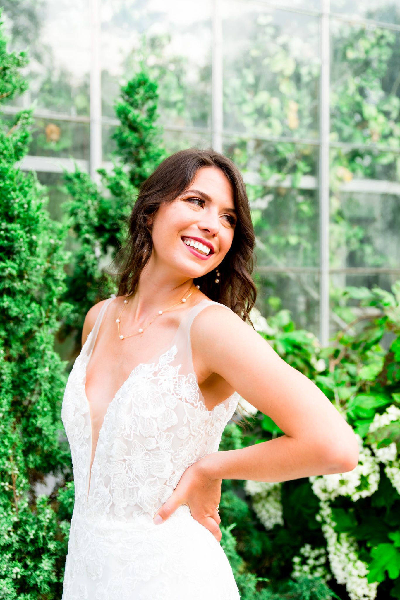 bride looking back and smiling with hands on hip in Serafina Gown and gold and pearl necklace at conservatory wedding