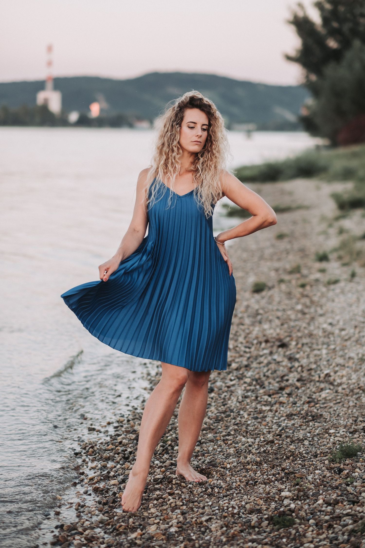 Person in blue pleated dress stands barefoot on rocky shoreline during sunset.