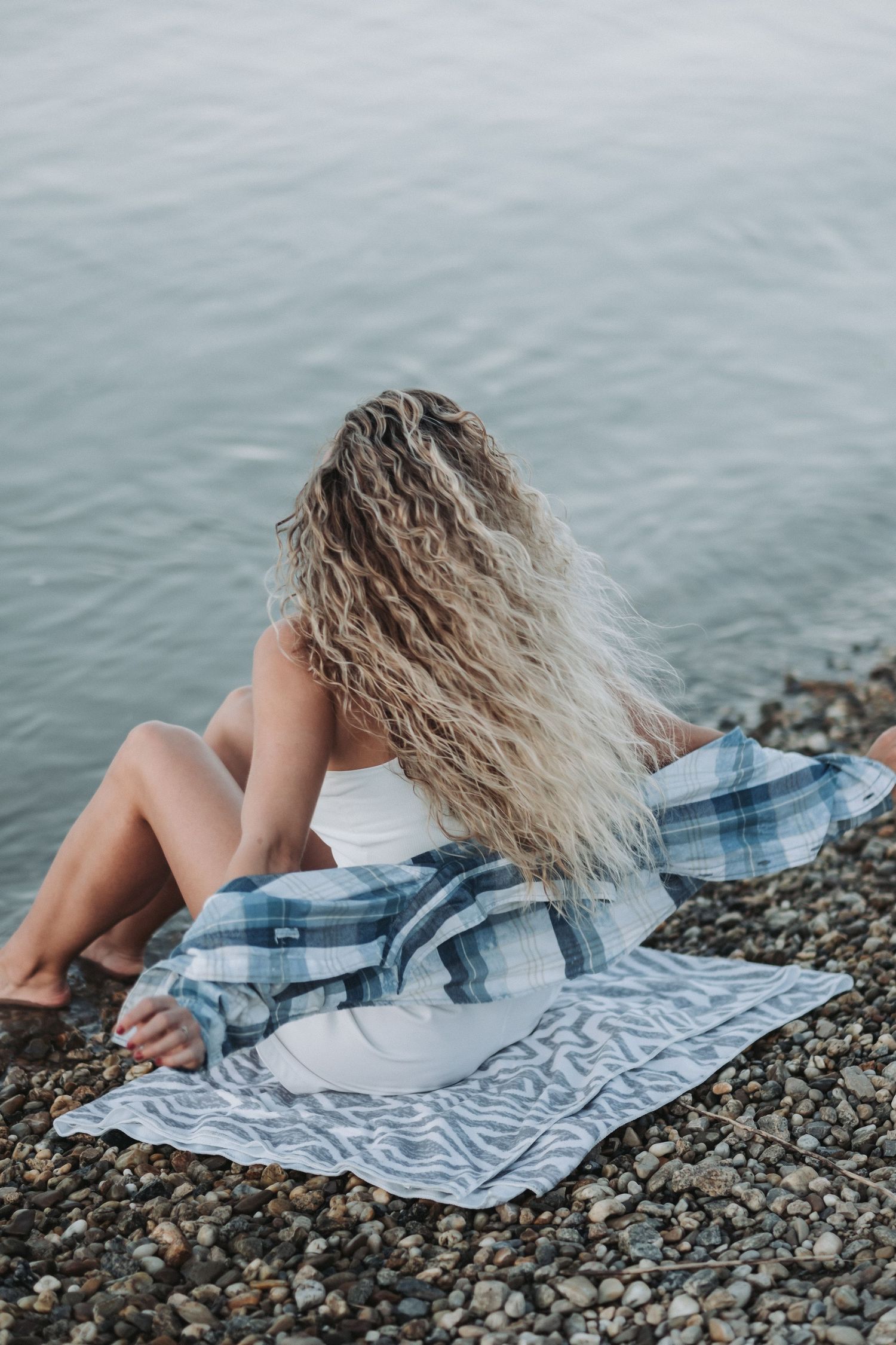 Woman with long wavy hair relaxing on rocky beach with plaid blanket at sunset.