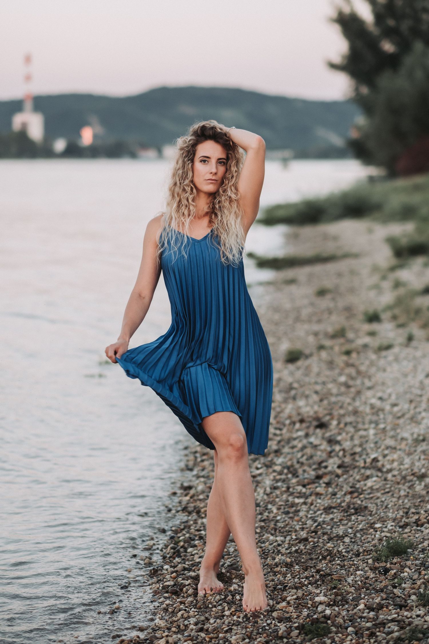 Figure in flowing blue dress poses on pebble beach with mountains in background.