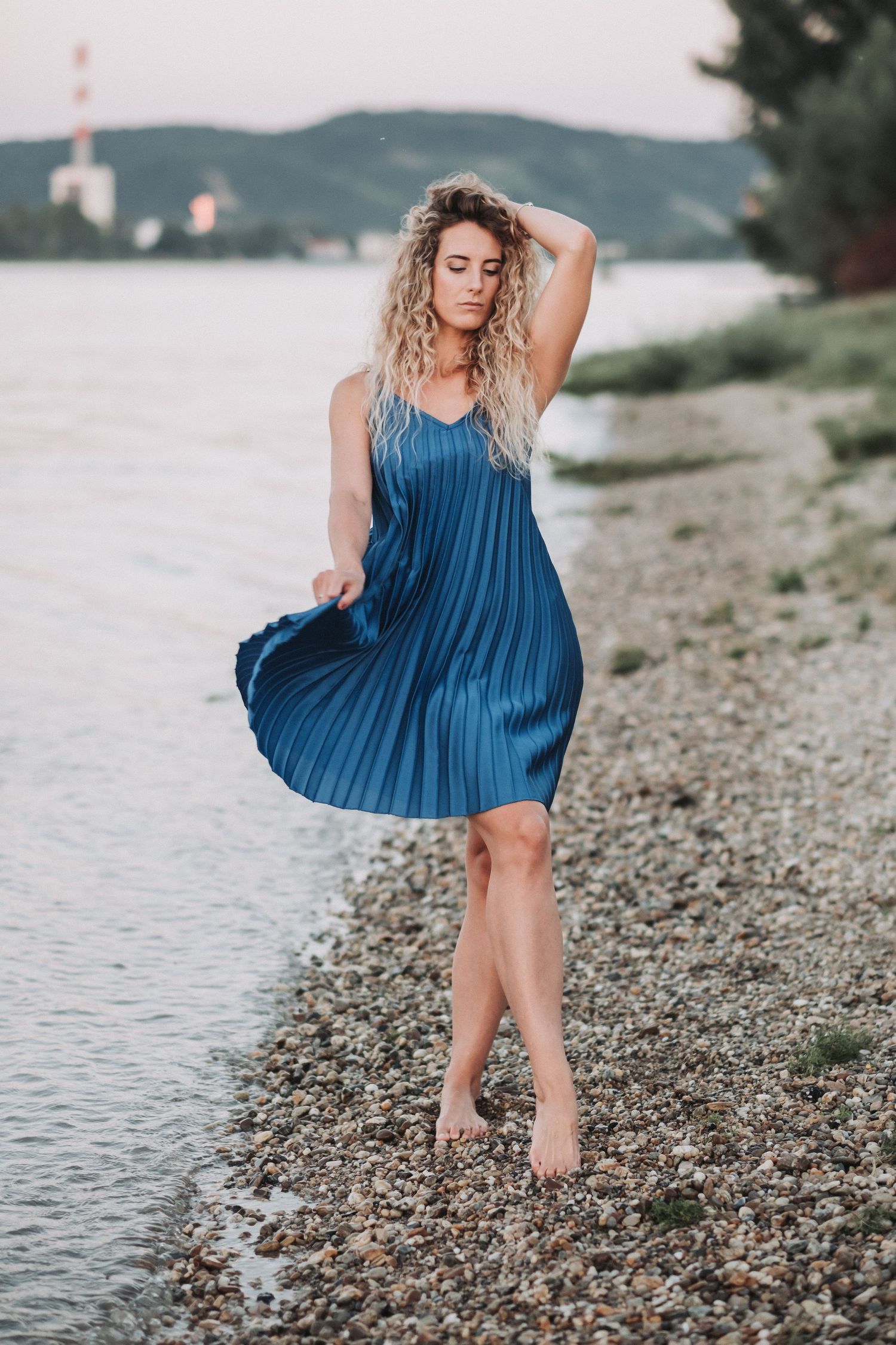 Person in blue pleated dress stands elegantly on rocky beach at dusk.