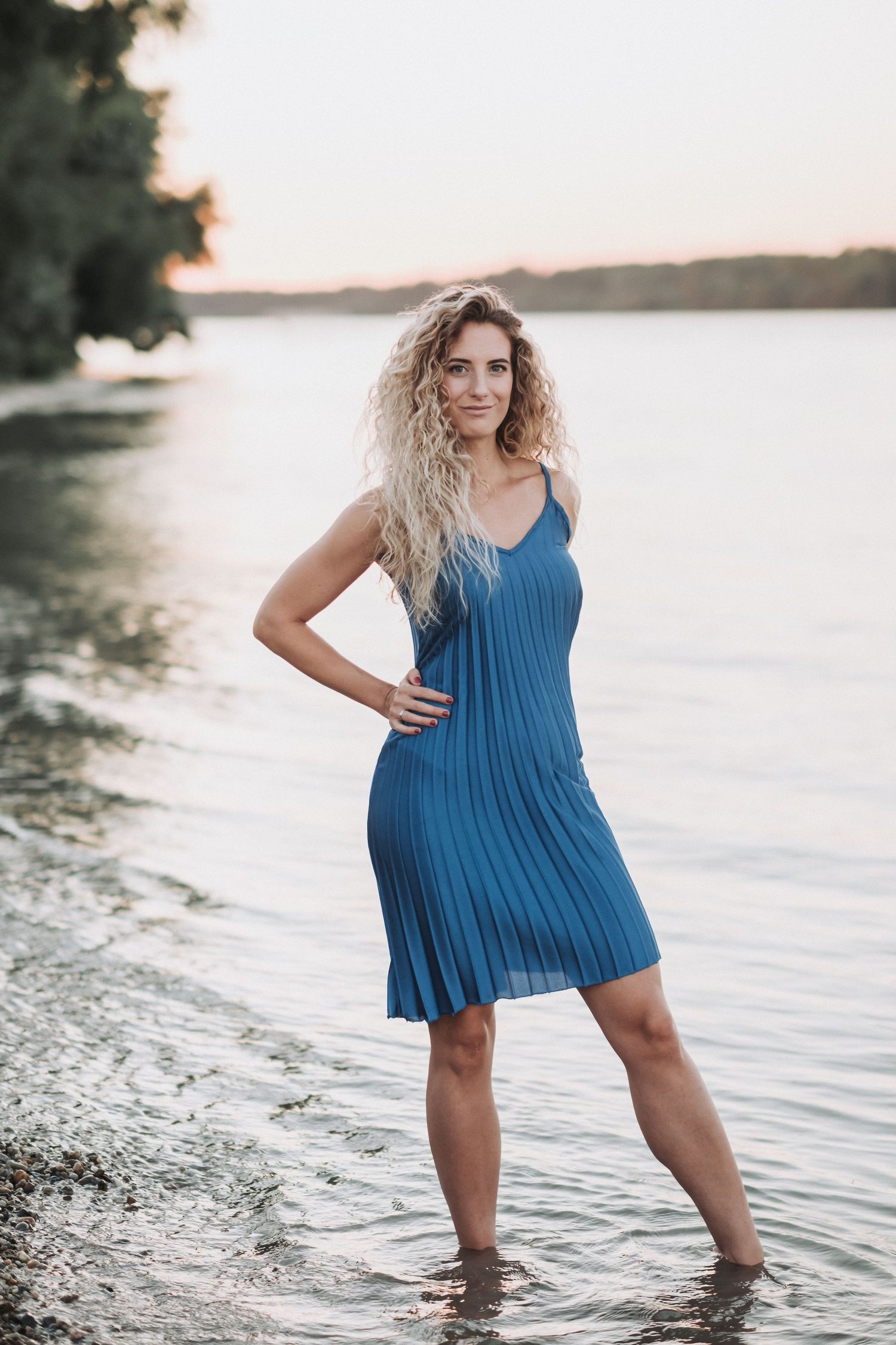 Figure in blue pleated dress stands in shallow water at rocky beach during sunset.