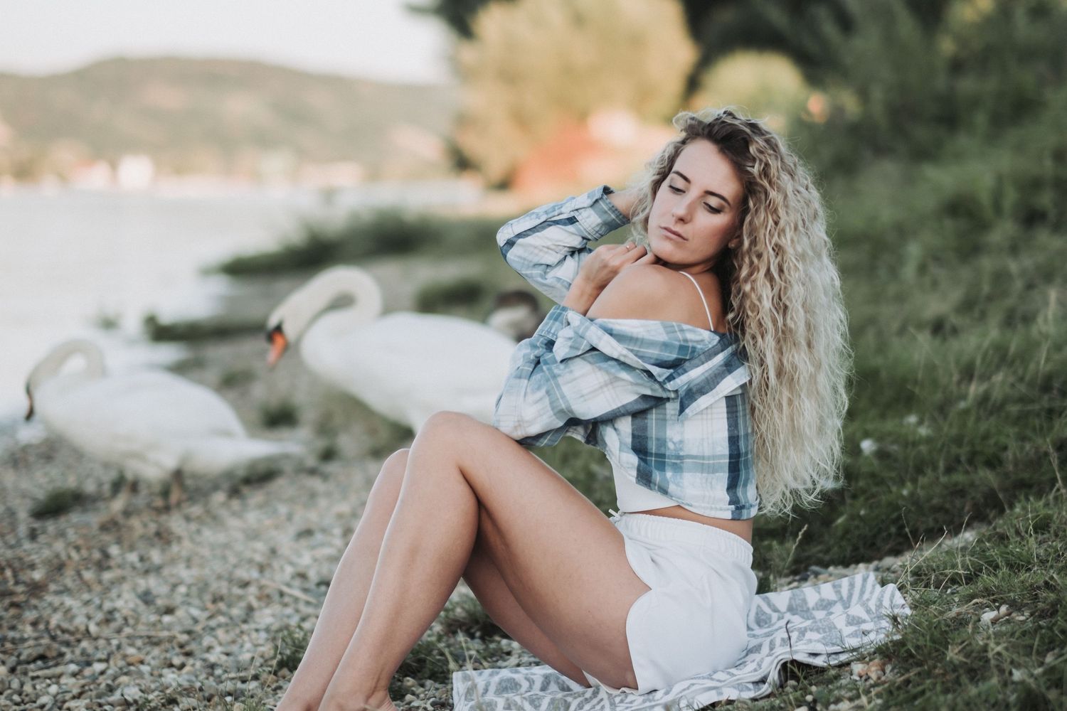 Model in plaid shirt and white shorts poses on rocky beach at sunset.
