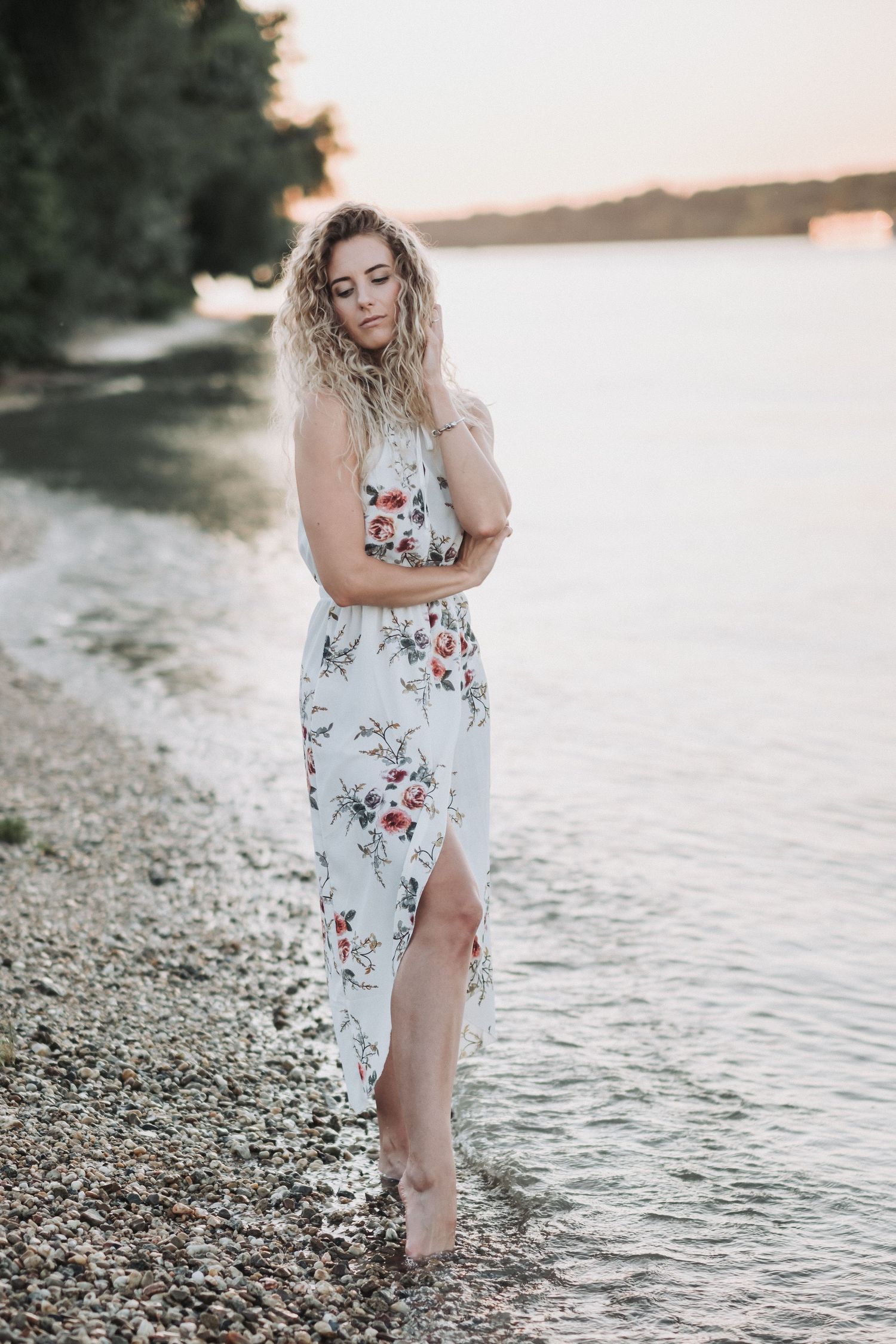 Person wearing white floral dress stands at water's edge during sunset.