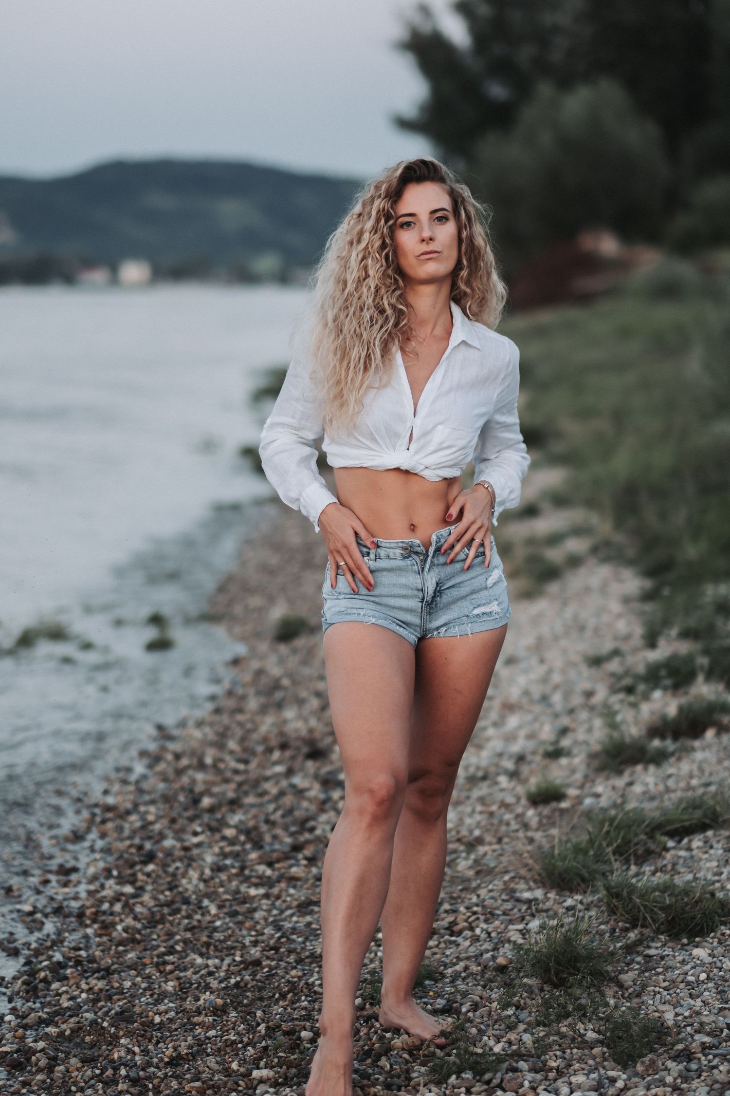 Fashion portrait of model in casual summer outfit standing barefoot on pebble beach.