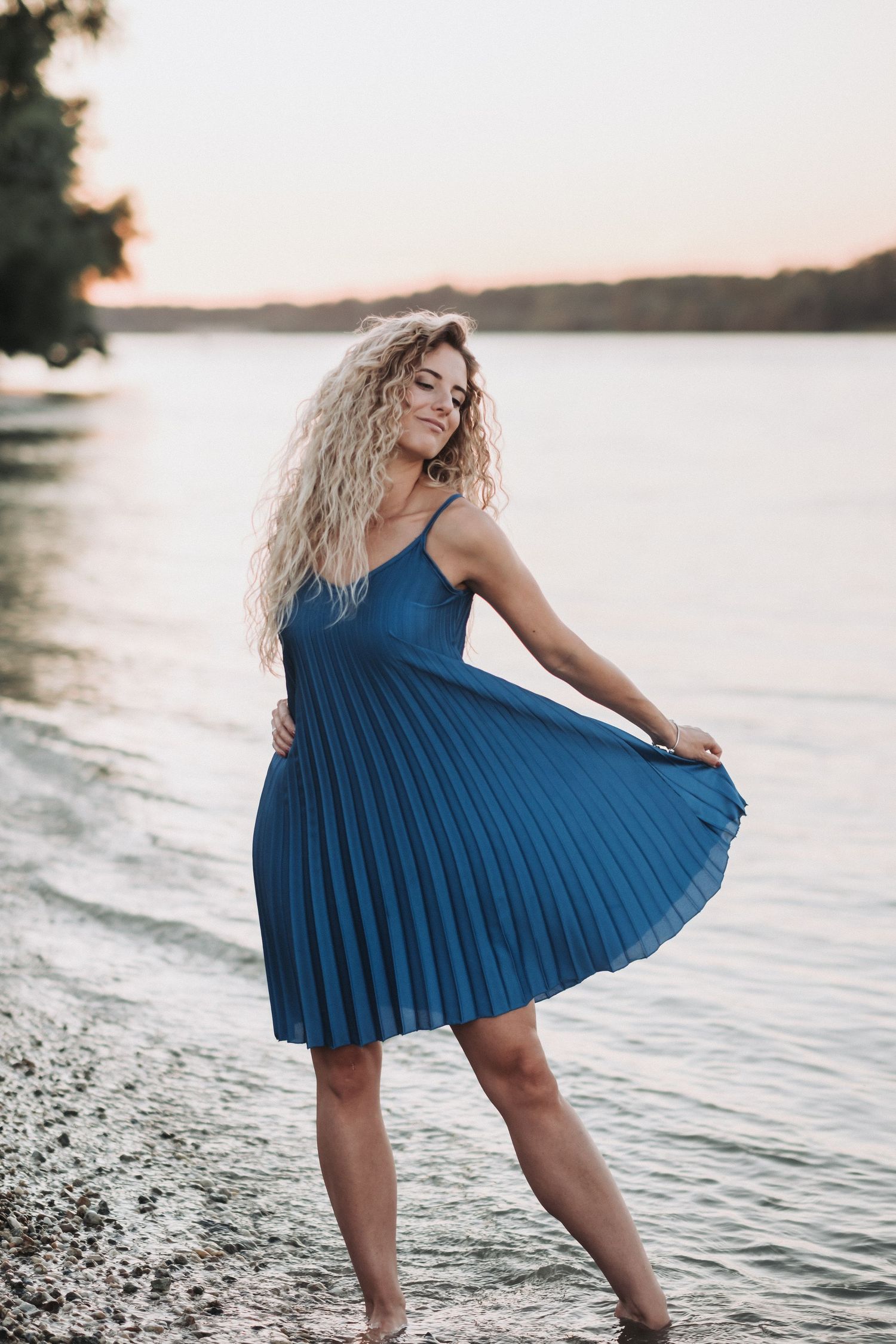 Woman twirls in blue pleated sundress on pebble beach at dusk.