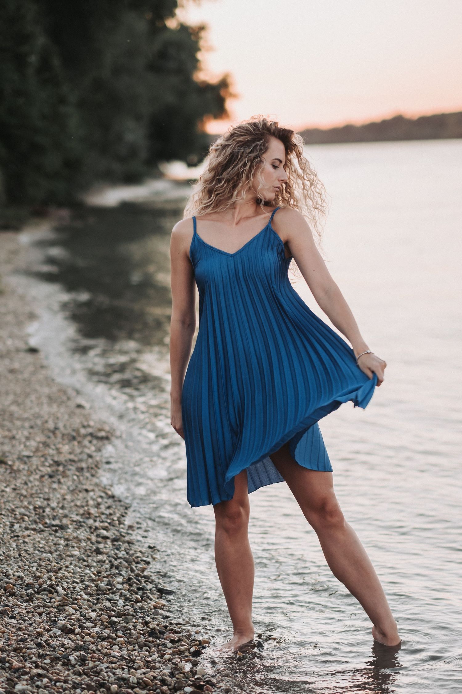 Person in flowing blue dress dances on rocky shoreline at sunset.