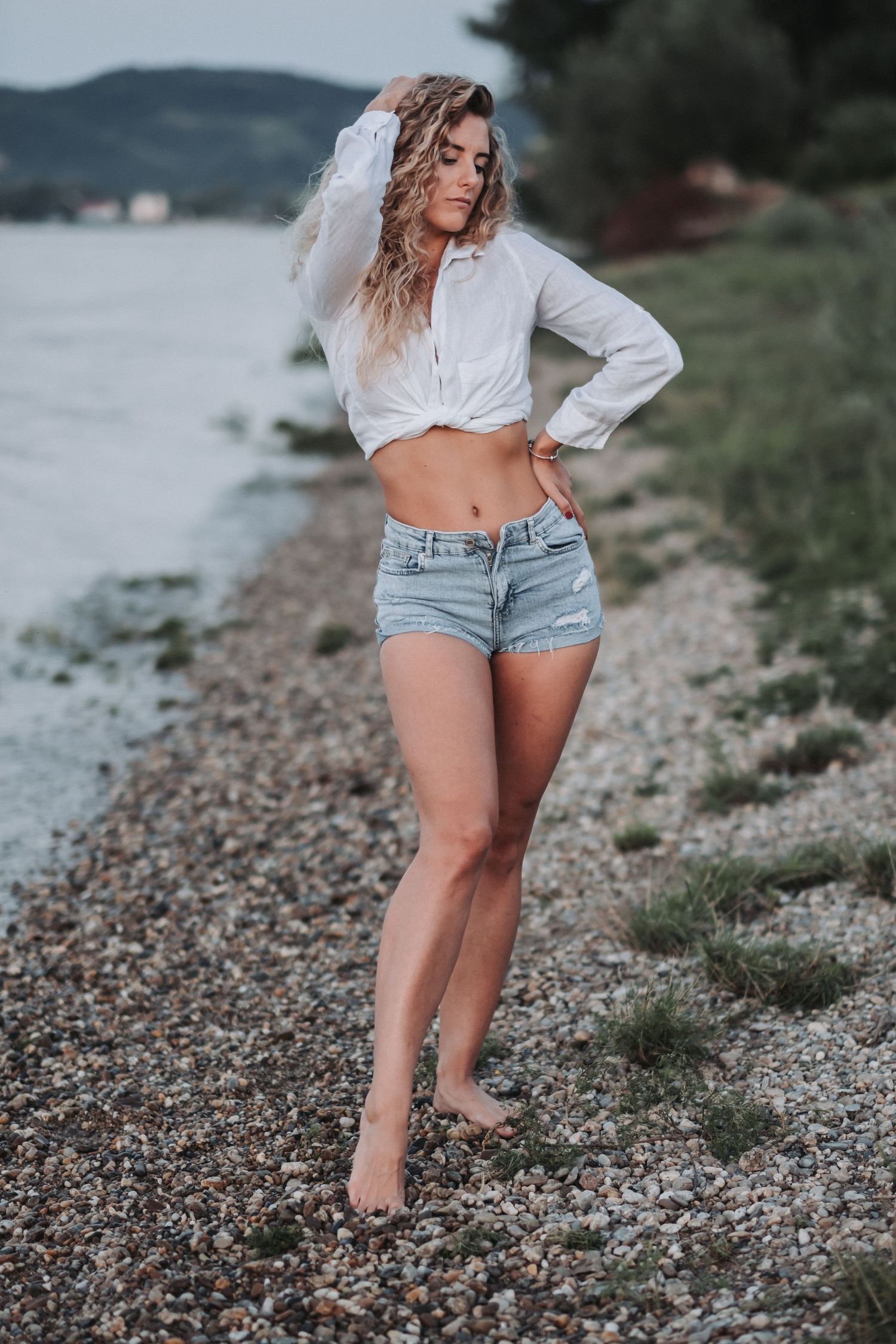 Casual beach photoshoot featuring white button-down shirt and jean shorts against lakeside backdrop.