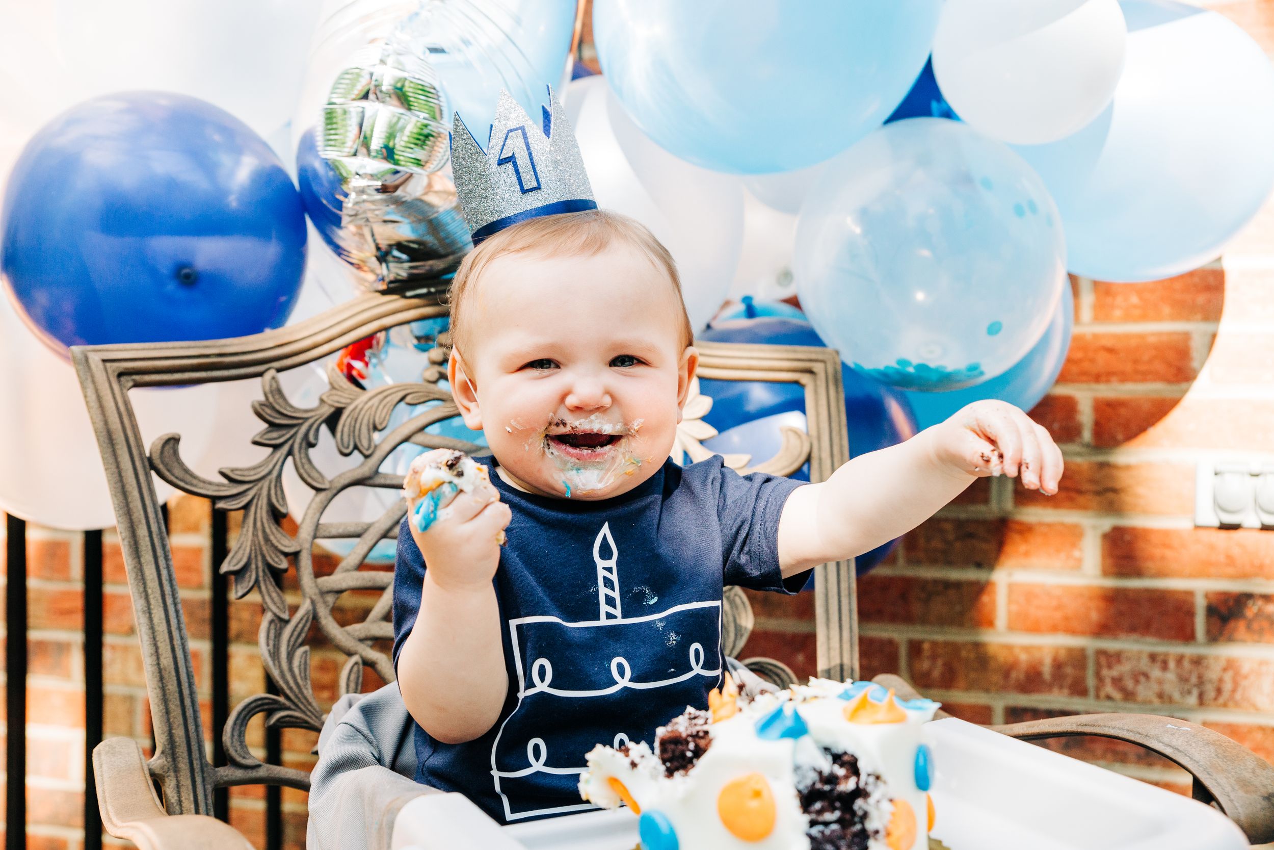 One year old boy surrounded by balloons and smiling while eating cake with his hands