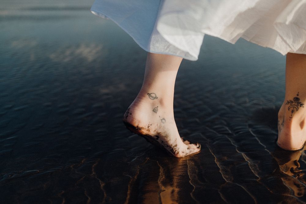 Bare feet with delicate gold temporary tattoos standing on wet reflective dark surface.