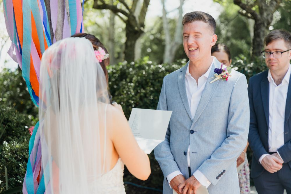 An outdoor wedding ceremony featuring bright blue and orange ribbon decorations and a light gray suited groom.