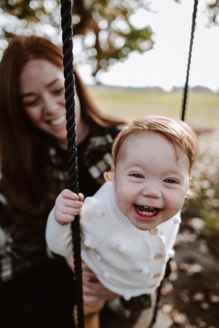 A joyful baby wearing a white cardigan laughs while being pushed on a rope swing outdoors.