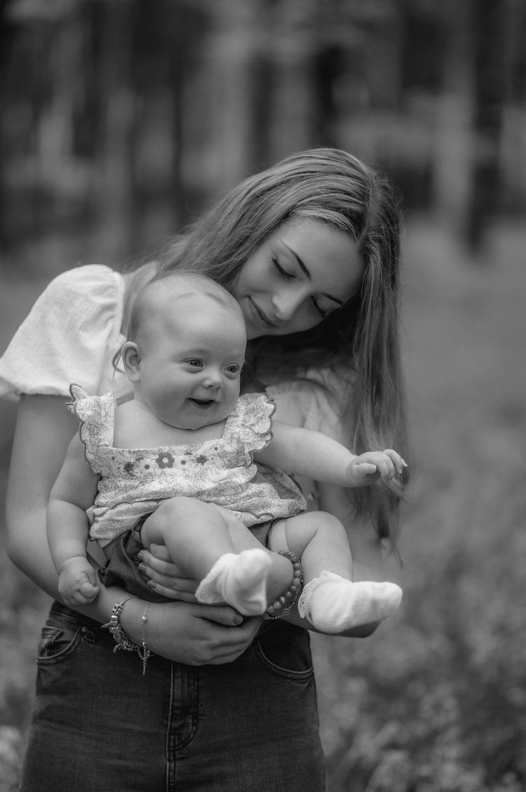 A tender black and white portrait of parent and baby cuddling outdoors in nature, baby wearing frilly outfit.