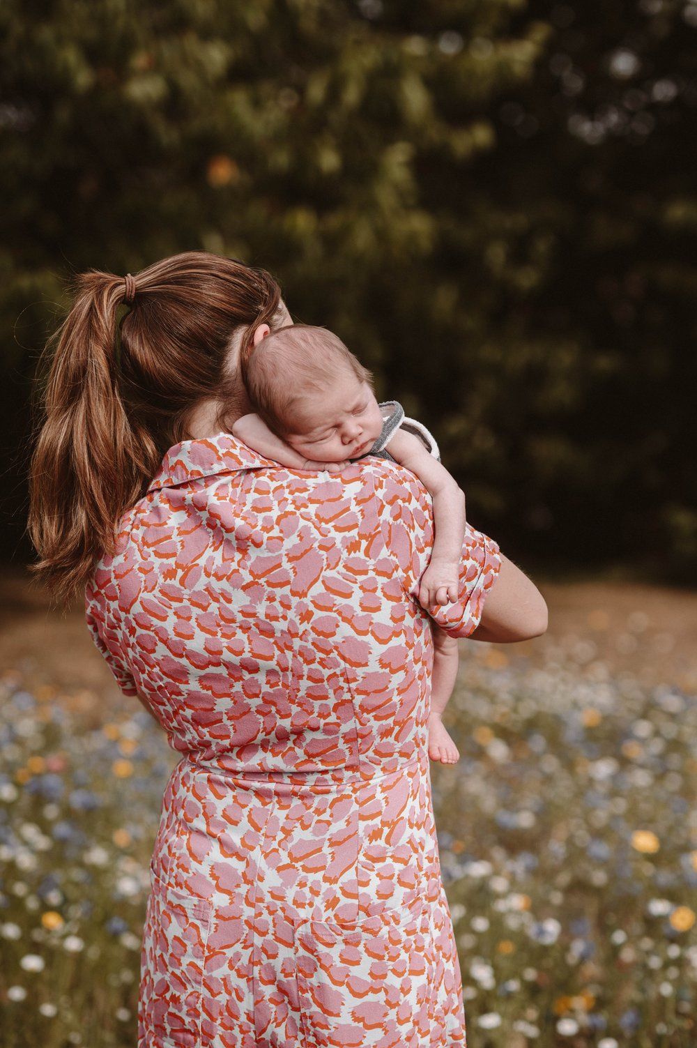 A person wearing a coral patterned dress holds a sleeping infant while standing in a wildflower meadow at sunset.