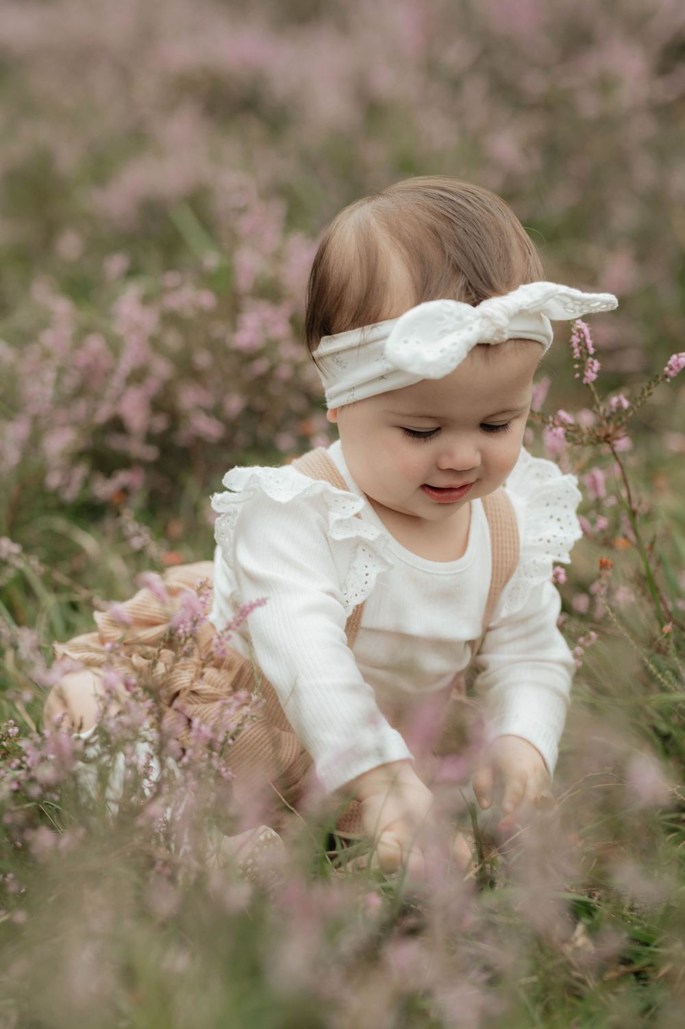 A baby wearing a white dress and bow headband sits among delicate pink wildflowers in a dreamy garden setting.