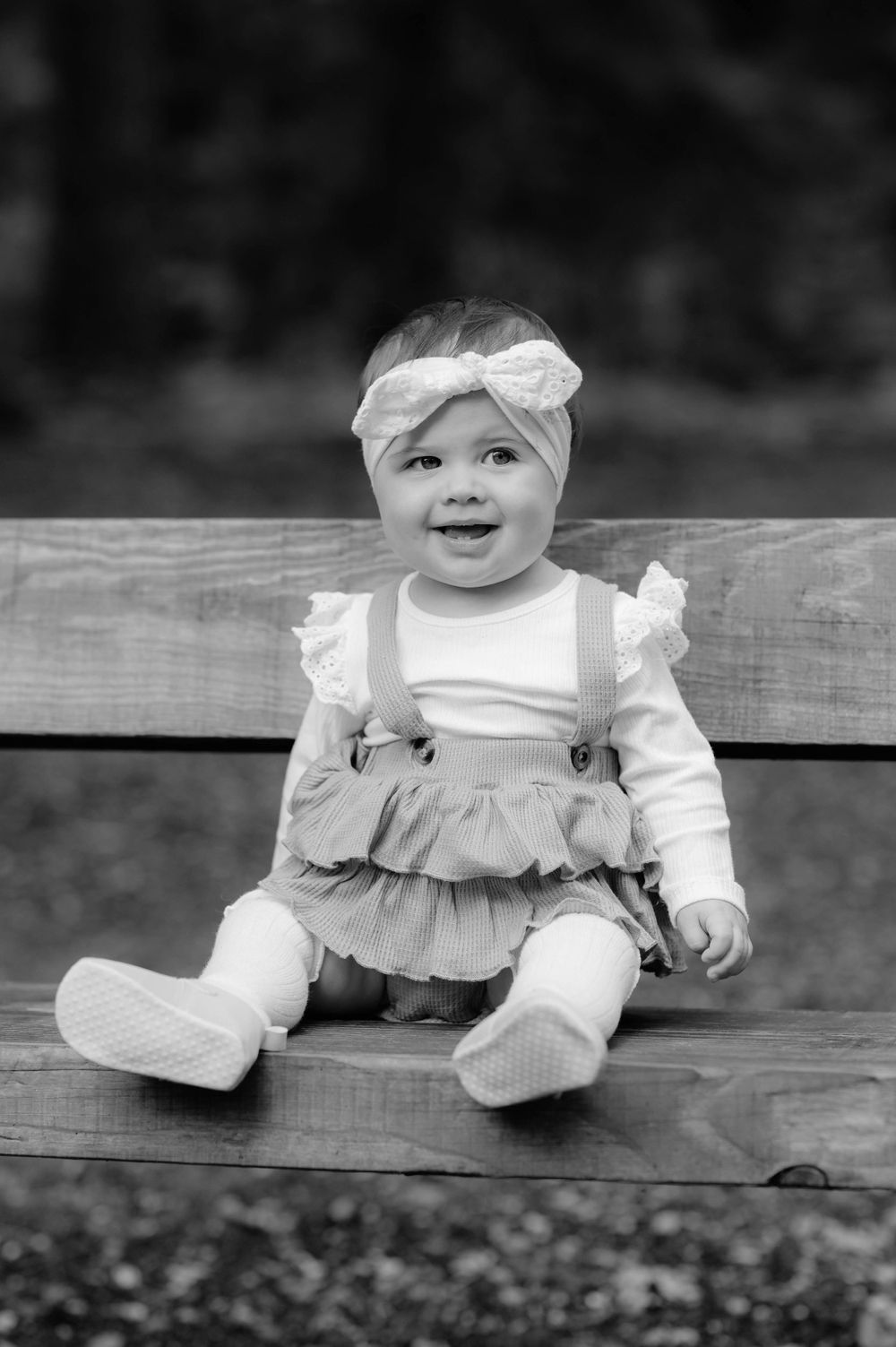 A black and white photo of a baby wearing a bow headband, ruffled dress, and overalls sitting on a wooden bench outdoors.