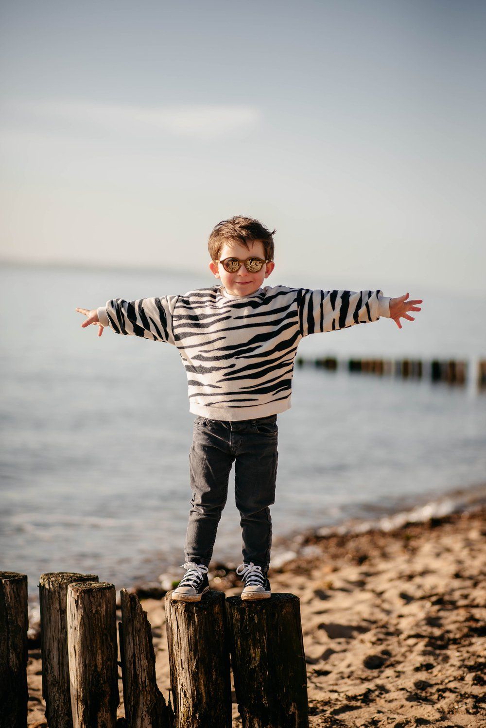 Young child in zebra striped sweater stands with arms outstretched on wooden post at beach during sunset.