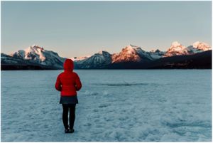 woman in red puffer jacket standing on frozen lake McDonald in glacier national park during the winter at alpenglow
