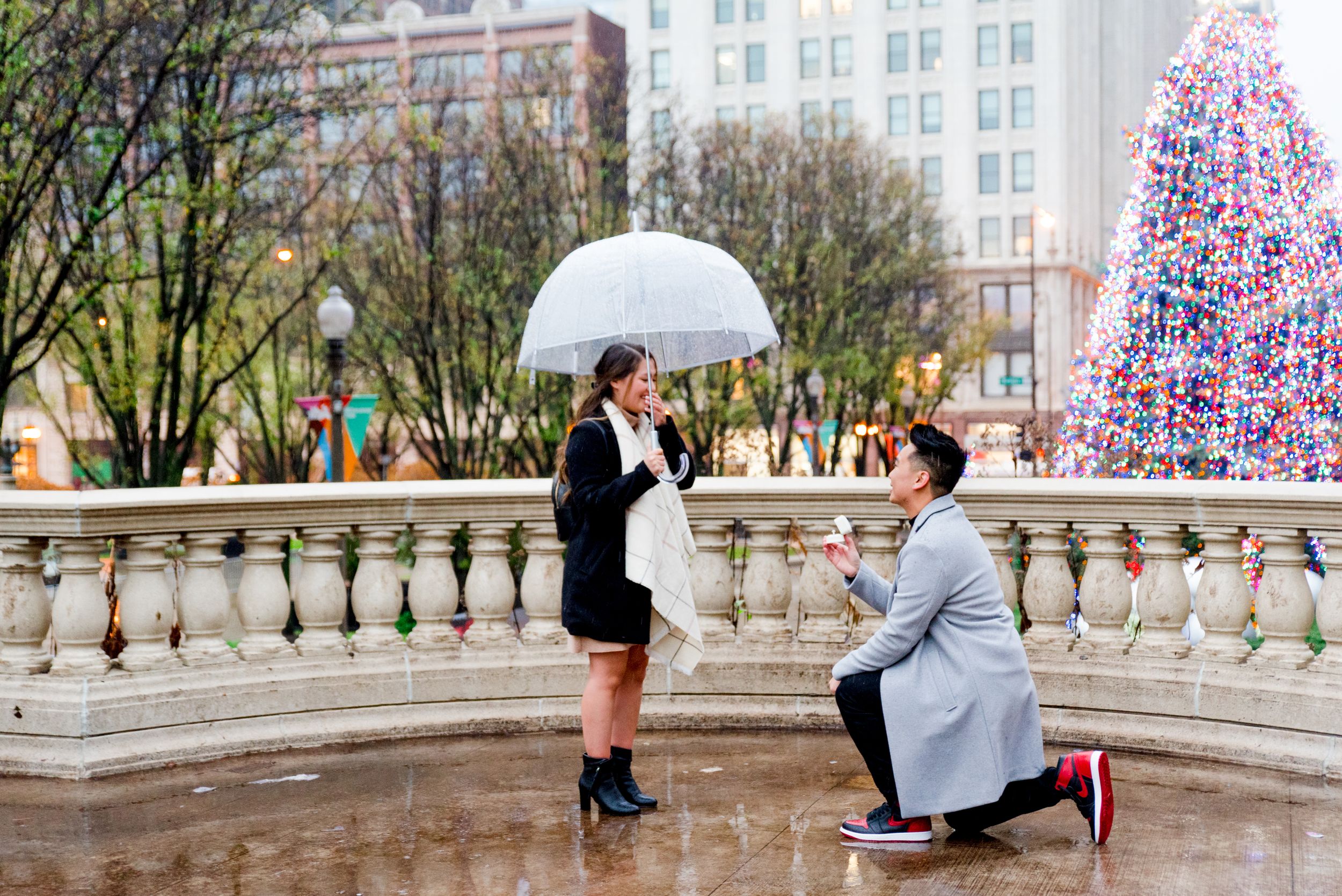 man in grey coat kneeling proposing to woman in black coat and clear umbrella in front of Millennium Park Christmas Tree