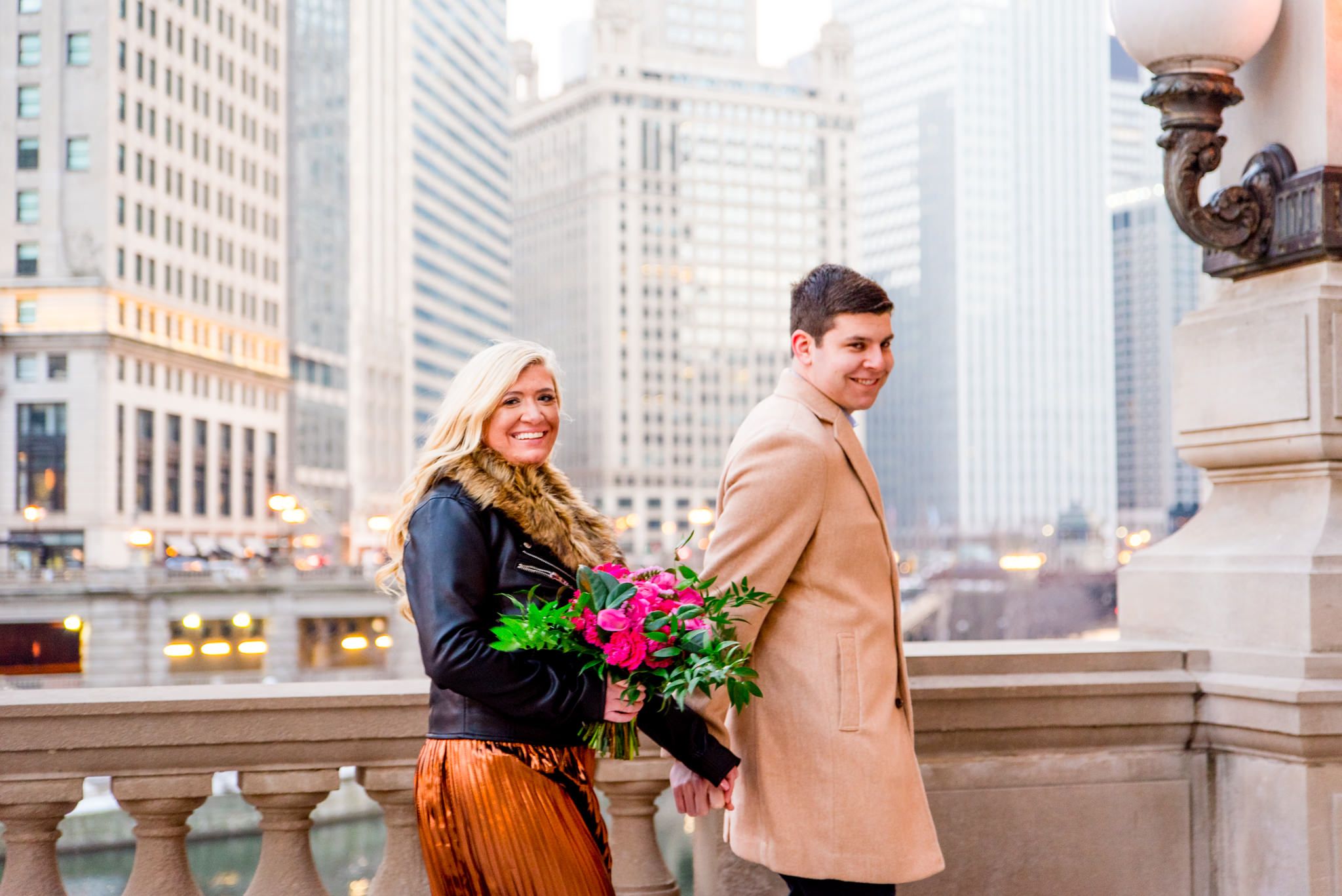 man in tan coat walking with woman in copper skirt in front of Wrigley Building engagement photos