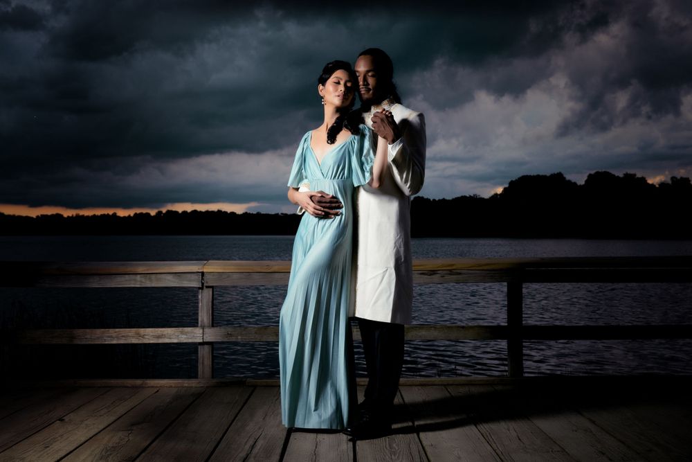 Romantic couple in formal wear embrace on dock at sunset with dramatic stormy sky backdrop.