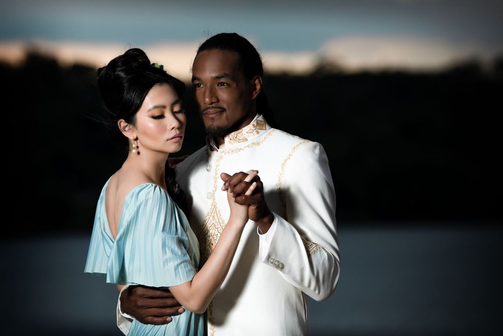 Elegant couple in white and blue formal wear dance together on waterfront deck at dusk.