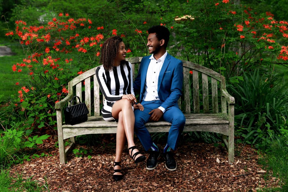 A romantic couple sits together on a wooden bench in a garden with vibrant orange flowers blooming in the background.