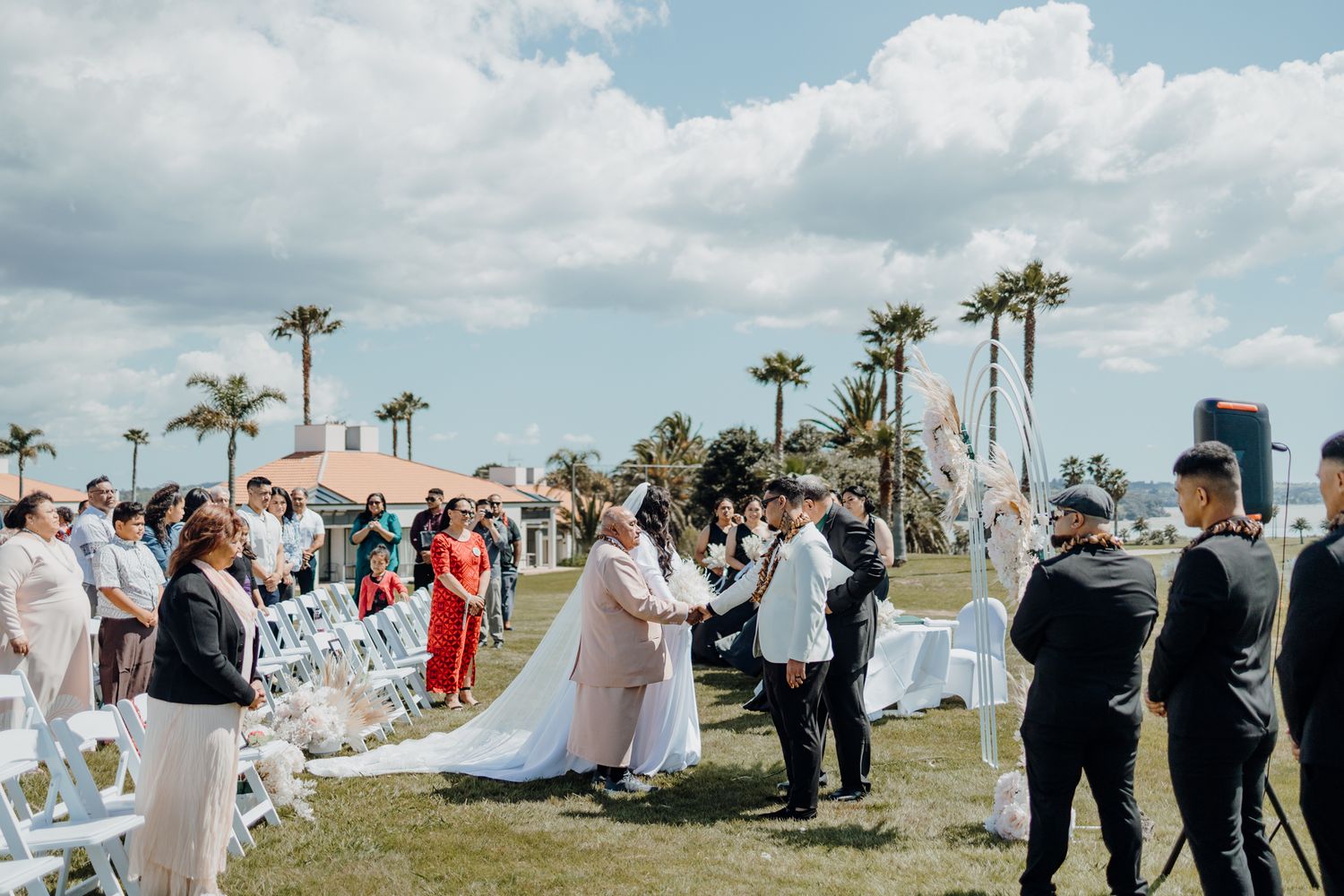 Outdoor wedding ceremony with palm trees and white chairs set up on a grassy lawn by the ocean.