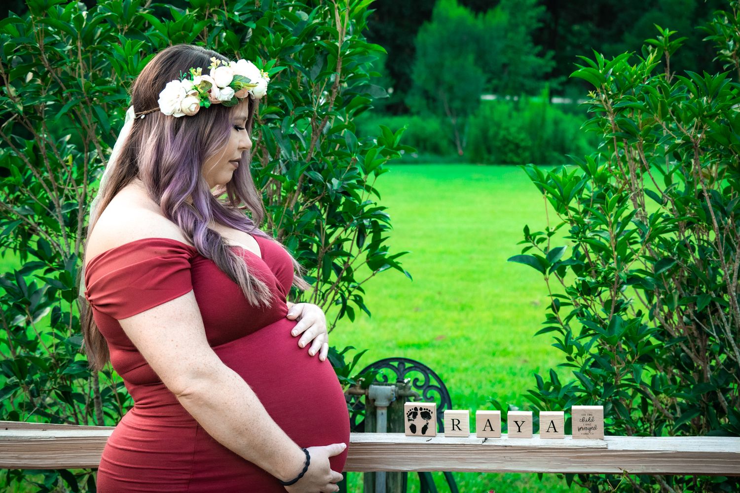 Pregnant woman wearing a red dress and flower crown stands in a garden with baby blocks on a ledge. Cherokee Park, Thomasville, Georgia