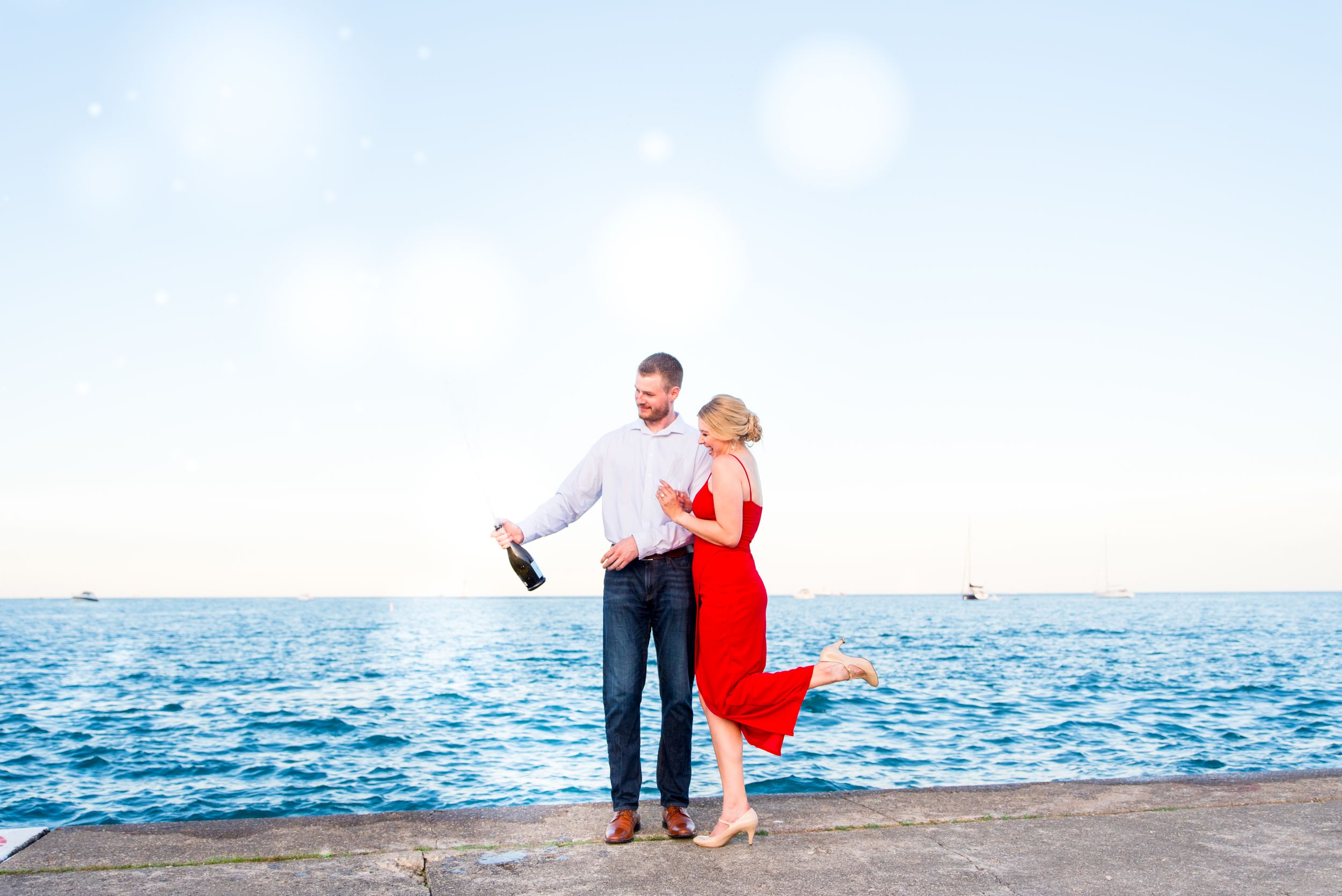 man pops bottle of champagne while blonde woman in red dress holds his arms and smiles by Lake Michigan